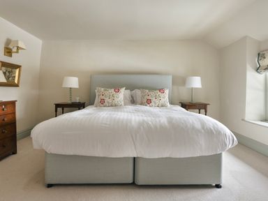 A bedroom with a double bed with white bedding and floral pillows two side tables with lamps a window and a wooden chest of drawers at High Lindeth in Bowness