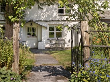 A pathway leading to a white cottage entrance with a table and two chairs outside surrounded by plants and flowers at High Lindeth in Bowness