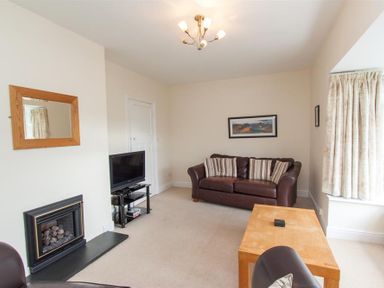 A living room with a brown leather sofa striped cushions a wooden coffee table a television on a stand a fireplace with a mirror above and cream curtains at The Haven in Keswick