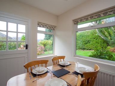 A dining area with a round wooden table and four chairs set with white plates and glasses by windows with views of a garden at The Haven in Keswick