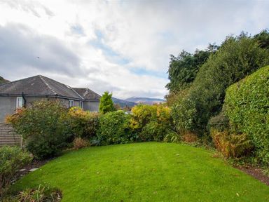 A garden with green grass and various shrubs and trees with houses and mountains in the background at The Haven in Keswick