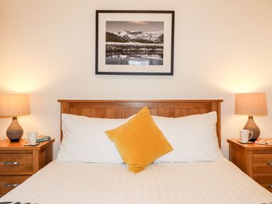 A bed with white pillows and a yellow cushion between two wooden bedside tables with lamps and cups at Fir Howe in Skelwith