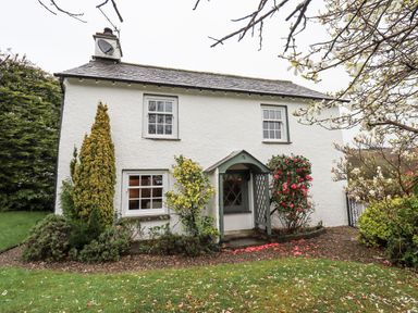 A white house with a slate roof and a small porch surrounded by shrubs and trees at Syke Cottage in Hawkshead