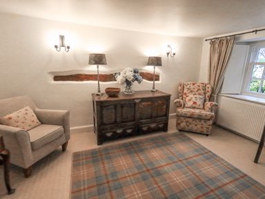 A living room with two armchairs a wooden sideboard with lamps and flowers and a window with curtains at Syke Cottage in Hawkshead