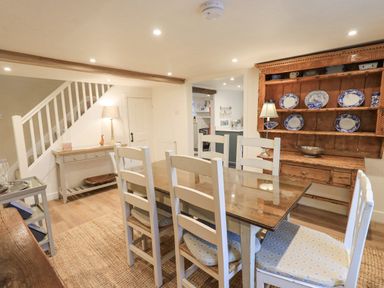 A dining room with a wooden table and white chairs a wooden dresser with blue and white plates a side table and stairs at Syke Cottage in Hawkshead