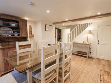 A dining room with a wooden table and white chairs a sideboard a lamp and stairs at Syke Cottage in Hawkshead