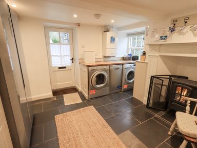 A utility room with washing machine dryer dishwasher a wood burning stove chair rugs and a door at Syke Cottage in Hawkshead