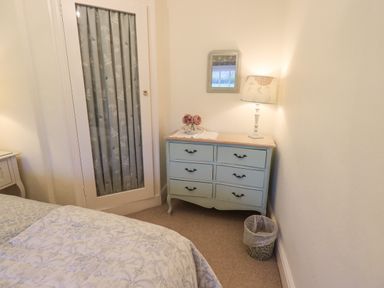 A bedroom corner with a blue dresser a table lamp a small mirror flowers and a waste bin at Syke Cottage in Hawkshead