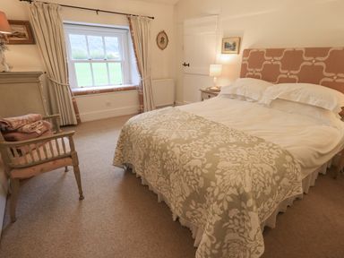 A bedroom with a bed with patterned headboard and bedspread next to a window with curtains and a chair with folded towels at Syke Cottage in Hawkshead