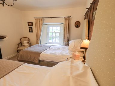A bedroom with two single beds, a window with curtains, an armchair with towels, and a bedside lamp at Syke Cottage in Hawkshead