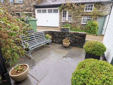 A small stone patio area with a metal bench potted plants and a stone wall in front of a stone building with white garage doors at Syke Cottage in Hawkshead