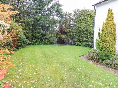 A grass garden with trees shrubs and two chairs with a small table at Syke Cottage Hawkshead