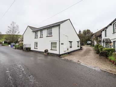 A white painted cottage with green trim on windows and a gravel driveway beside a road at Syke Cottage in Hawkshead