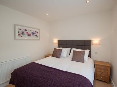 A bedroom with a bed with purple cushions and blanket two wooden bedside tables and floral artwork on the wall at Mylnbeck in Bowness