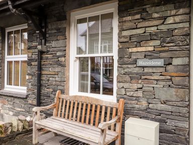 A stone wall exterior with two white framed windows a wooden bench and a sign reading Hazeldene in Ambleside