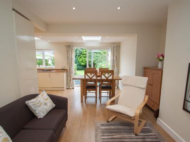 A living room and dining area with a wooden dining table and chairs a white cushioned chair sofa with cushions wooden cabinet and kitchen sink at Fell End Keswick