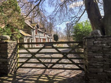 A stone cottage behind a wooden gate with a gravel driveway and trees at The Stables in Ambleside