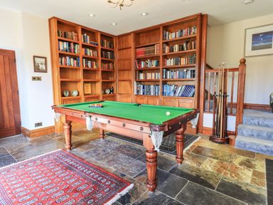 A game room with a pool table, wooden bookshelves filled with books, a red patterned rug, and a staircase at The Stables in Ambleside