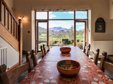 A dining room with a long table and wooden chairs overlooking a garden and mountains through large windows at The Stables Ambleside