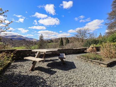 An outdoor area with a wooden picnic table on gravel surrounded by low stone walls and plants with hills and trees in the background at The Stables in Ambleside