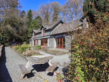 A stone house with wooden framed windows and a gravel pathway with a wooden picnic table outside surrounded by trees and bushes at The Stables in Ambleside