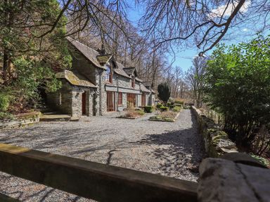 A stone house with gravel driveway and garden beds surrounded by trees at The Stables in Ambleside