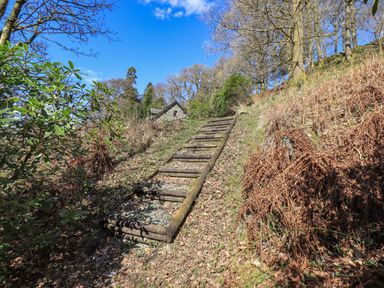 An outdoor wooden log stairway on a forest slope with leaf covered ground and a stone building in the background at The Stables in Ambleside