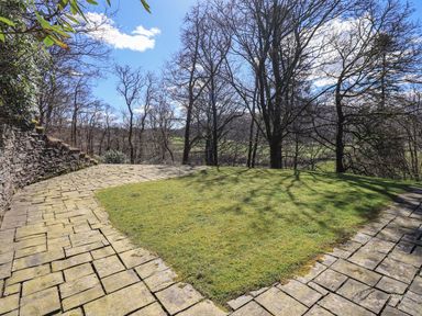 An outdoor area with a paved stone path surrounding a grass patch and trees in the background at The Stables in Ambleside