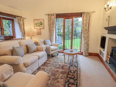 A living room with beige sofas a patterned rug a glass coffee table fireplace and large windows with curtains at Hannakin Barn in Hawkshead
