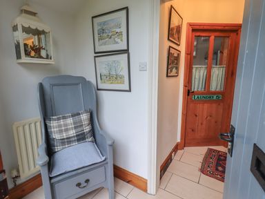 A hallway with a blue chair with cushions near framed pictures on the wall and a wooden door with a laundry sign at Hannakin Barn in Hawkshead
