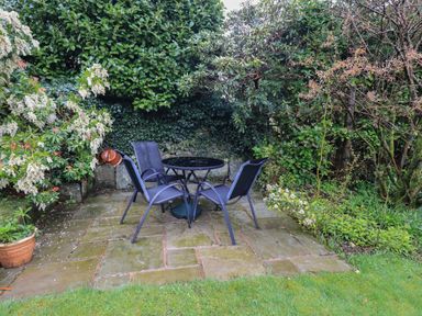 A garden patio with a black round table and three black chairs surrounded by green shrubs at Hannakin Barn in Hawkshead