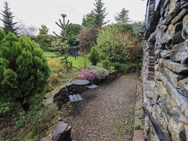A gravel pathway next to a stone wall with metal table and chairs and surrounding greenery at Hannakin Barn in Hawkshead