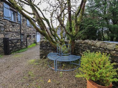 A stone house with blue window shutters a gravel pathway a circular metal bench around a mossy tree and a potted plant at Hannakin Barn in Hawkshead