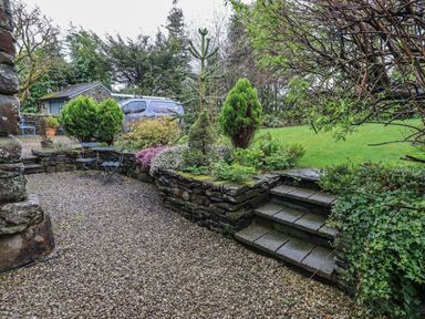 An outdoor garden area with stone steps, gravel ground, small trees, shrubs, and a stone wall with a small shed and a parked van in the background at Hannakin Barn in Hawkshead