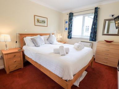 A bedroom with a wooden bed and white bedding with pillows and towels on the bed a wooden nightstand with a lamp a window with blue curtains and a wooden dresser with a mirror and television on the wall at Fairview in Bowness