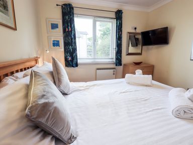 A bedroom with a wooden bed with white bedding and pillows a window with blue curtains a wooden dresser with a red bowl and a wall-mounted TV at Fairview in Bowness