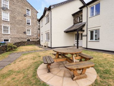 A wooden picnic table on a circular paved area in a grass yard next to a white building with multiple windows Kerris Place Fairview Bowness