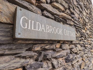 A stone wall with a rectangular sign that reads Gildabrook Cottage in Bowness