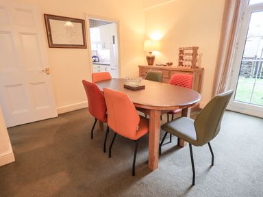 A dining room with a wooden oval table surrounded by six multicolored chairs a cabinet with a lamp and wine rack and a door leading to a kitchen at Gildabrook Cottage in Bowness