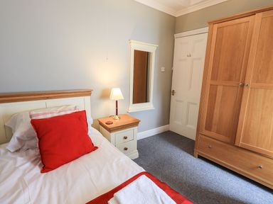 A bedroom with a single bed with red cushions a bedside table with a lamp a wall mirror a wardrobe and a closed door at Gildabrook Cottage in Bowness