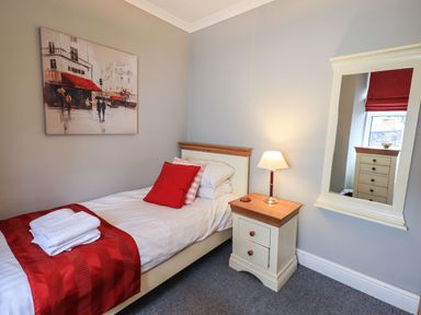 A single bed with red pillows and a red runner next to a bedside table with a lamp and a wall mirror in a bedroom at Gildabrook Cottage in Bowness
