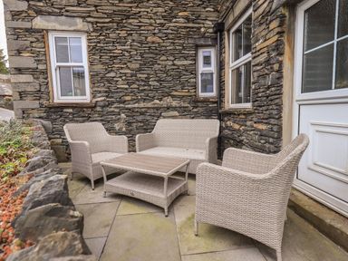 An outdoor seating area with a wicker sofa two wicker chairs and a wicker coffee table against a stone wall at Gildabrook Cottage in Bowness