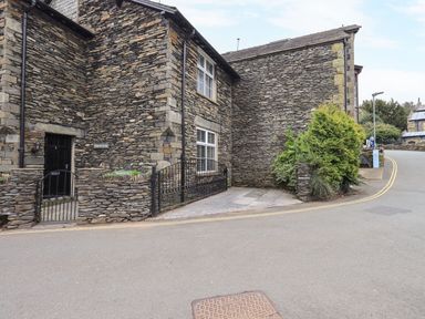 The exterior of a stone building with two windows a black door a small gated area and some greenery beside a road at Gildabrook Cottage in Bowness