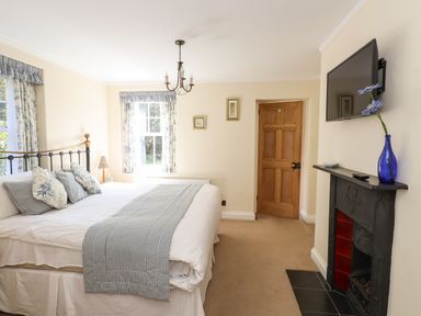 A bedroom with a double bed pillows and blanket two windows wooden door wall-mounted TV and a fireplace with flower vase at Hodge How Cottage in Windermere