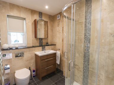 A bathroom with a shower enclosure, a toilet, a sink with a wooden cabinet, and a small window at Hodge How Cottage in Windermere