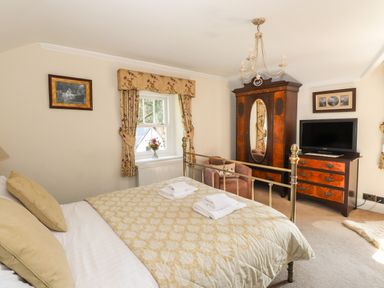 A bedroom with a bed a window with curtains an armchair a wardrobe a dresser with a television and framed pictures at Hodge How Cottage in Windermere
