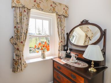 A window with floral curtains and a vase of orange flowers next to a wooden dresser with an oval mirror and decorative items at Hodge How Cottage in Windermere
