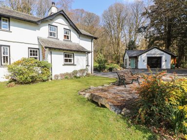 A white cottage with a garden bench and flowers near a detached garage at Hodge How Cottage in Windermere