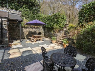 An outdoor patio with a wooden picnic table and umbrella stone steps and metal table and chairs surrounded by greenery at Hodge How Cottage in Windermere