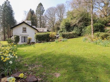 A white house with black trim surrounded by green lawn trees and yellow flowers at Hodge How Cottage in Windermere